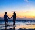 A romantic couple walks hand in hand on a tropical beach at sunset, enjoying a serene moment together.
